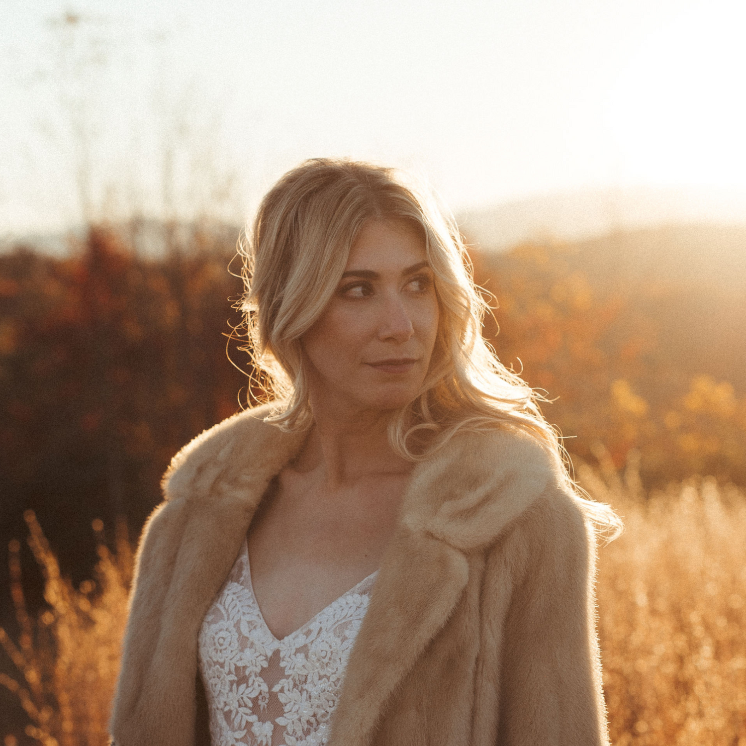 a woman in a lace top and fur coat, standing in a field during sunset. Her gaze is directed away from the camera, and the warm backlight from the sun enhances the autumnal tones of the surrounding landscape.
