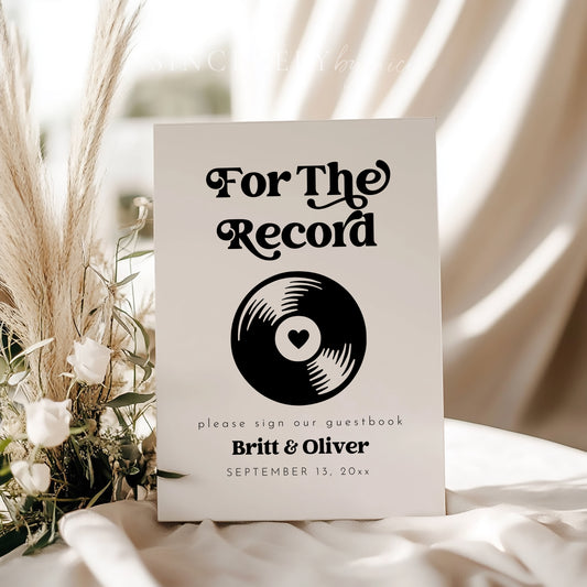 Minimalist retro-style guestbook sign reading "For The Record" with a vinyl graphic, styled with white roses and pampas grass for a wedding display.