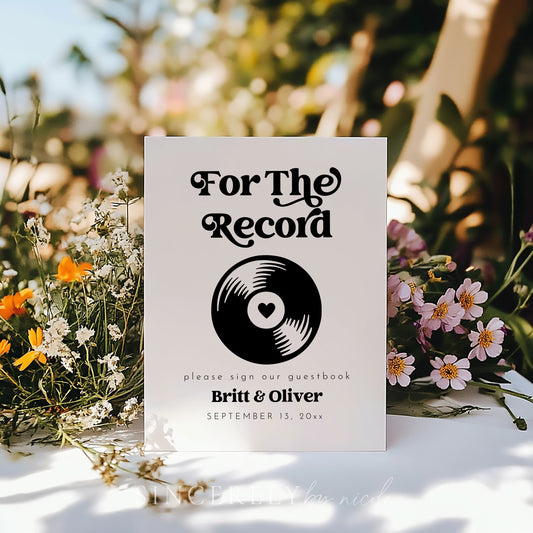 Modern vinyl record guestbook sign with bold typography surrounded by wildflowers in a sunny garden setting for an outdoor wedding.